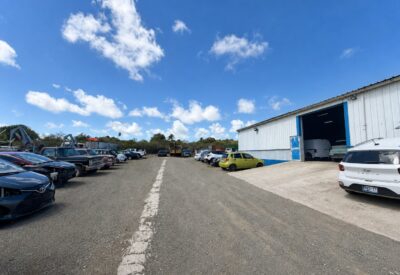 Parkeerplaats met diverse auto's en een grote loods onder een blauwe hemel.